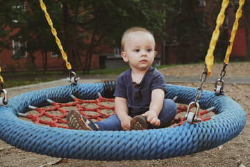 Pensive toddler boy swings on a round swing on playground. Baby spends time on playground.