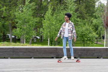 Skateboarding and urban lifestyle: trendy african american female riding longboard dressed in city style clothes. Black young woman skate boarding in city park. Freedom and carefree leisure concept