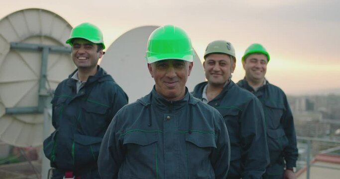 Portrait of group of professional telecommunication industry engineers smiles and looks at the camera . Workers wearing safety uniform and hard hat on unfocused background with sky and antennas .