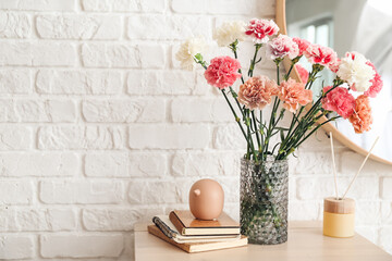 Vase with carnation flowers, books and reed diffuser on shelf near brick wall