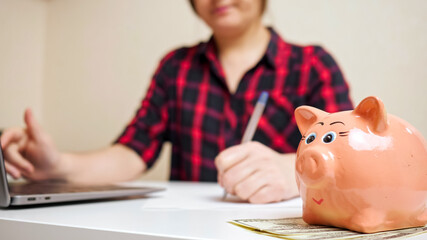 Obraz na płótnie Canvas Nice pink piggy bank stands on dollar banknotes against blurred lady typing on laptop keyboard and counting child expenses close view