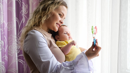Happy young mother with curly loose hair holds in arms baby girl in yellow bodysuit kissing near window with colorful curtains in apartment room