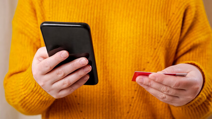 Young woman in stylish yellow sweater hands hold red credit card and enter information on black smartphone for online shopping close view
