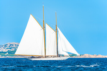 Fototapeta premium Stunning view of a wooden sailboat with white sails sailing on a blue water during a sunny day. Isola di Spargi, Maddalena Archipelago,Sardinia, Italy.
