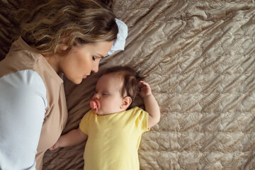 Caring young mother with curly hair lies on large bed near sleeping with pacifier baby girl and kisses gently in apartment room close upper view