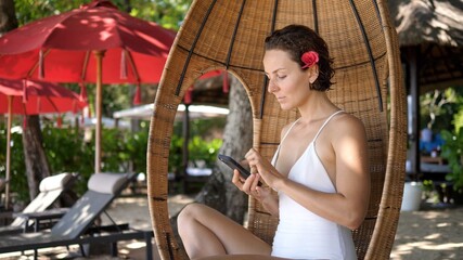 Beautiful young Caucasian woman in a white swimsuit and a red flower in her hair using her...