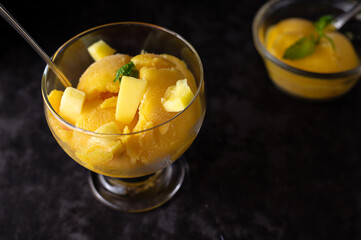 Mango yellow ice cream with fruit pieces and mint in glass. Horizontal, top view on a dark table