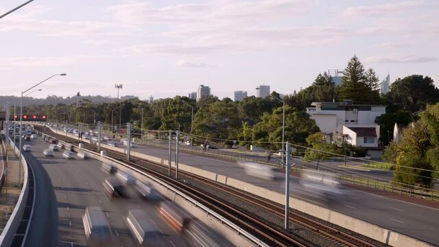 Busy traffic rushes along Perth freeway. Passenger train shoots up middle. City skyline visible beyond trees in background. Light clouds. Overhead view