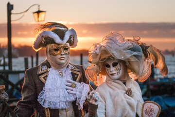 Male and female with masks, hats and carnival costumes standing by the water at sunset in Venice