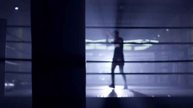 Kickboxer or Muay thai fighter training in low light gym . Silhouette on dark background . Boxer training in ring . Cinematic slow motion shot of a professional young man practicing in boxing ring