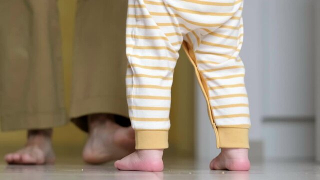 Baby boy taking first steps . Unrecognizable Little child girl walking on white floor at home. Barefoot legs mother and kid on kitchen