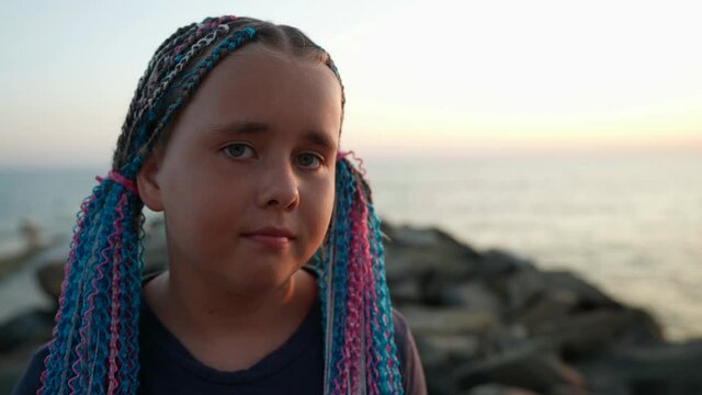 Portrait of a sad child a girl on the ocean shore in the setting sun multicolored pigtails are woven into her hair