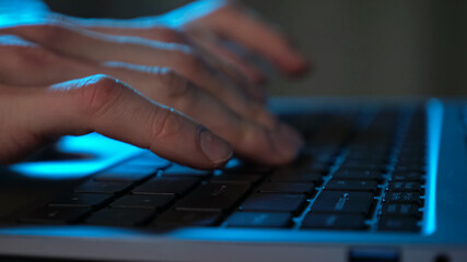 Close-up of man's hands typing on laptop keyboard in muted blue light.