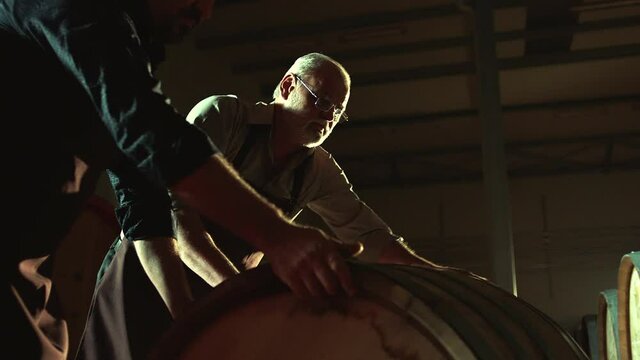 Worker in wine , whiskey or brandy warehouse sorting and rotating barrel . Two winemakers in vintage , traditional wine factory rolls barrel . Shot on ARRI ALEXA Cinema Camera in Slow Motion .