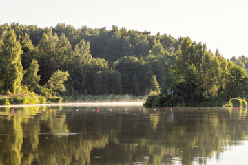 Dawn. The first rays of the sun illuminate the grass and trees on the river bank. Early morning, fog. Landscape with fog over water.