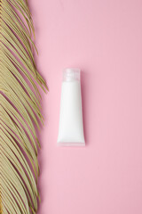 White tube of cosmetic cream with palm leaf on pink background. Flat lay