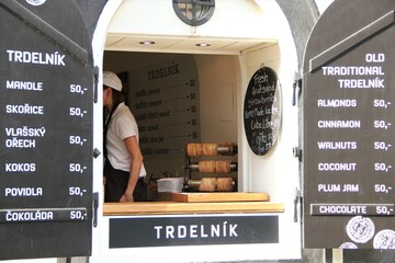 European sweet spit cake trdelník booth, a young female waitress inside, in the quaint Bohemian town of Český Krumlov with a list of available flavors on both sides