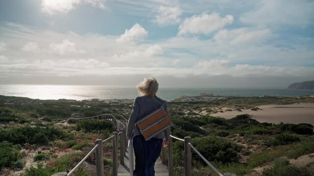 A middle-aged European woman artist with an easel on her shoulder walks towards the ocean.