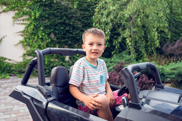 Excited little boy sits in a black electrocar in front of green fence and a tree. Holidays and free time for preschoolers