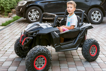 Fototapeta premium A bossy little boy sits in a black electrocar for kids in front of a big black car for adults. Child is driving
