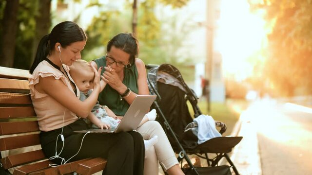 Multigenerational Family Uses A Laptop For Work In Nature By Spending Time Outdoors. Young Business Mother Holds A Baby In Her Lap And Uses A Laptop On A Park Bench