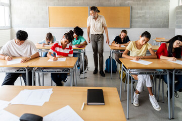 Mature adult caucasian man teacher walking in class to monitor multiracial group of students taking...