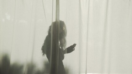 Silhouette of a long-haired girl standing behind curtains with her smartphone. Contrasting shadow in the bright sun. Summer mood. Young woman on vacation. Person texting messages and making phonecall.