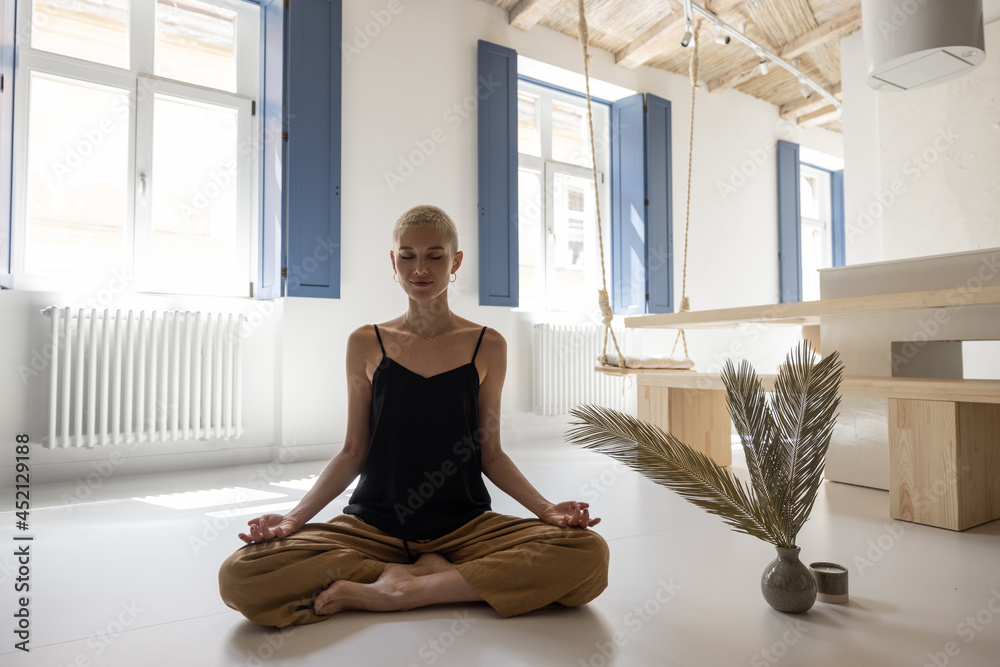 Wall mural Young stylish woman meditating and practicing yoga in a modern bright living room at home. Mindfulness, feeling calm at modern home