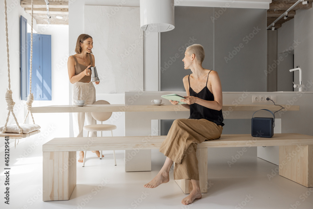 Wall mural Lesbian couple preparing breakfast, sitting with a digital tablet and drinking coffee at modern studio apartment. Concept of homosexual relationships and comfortable home