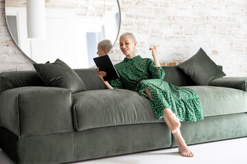 Stylish woman in green dress using digital tablet while sitting relaxed on the comfortable sofa at home. Leisure time with a digital device or working home concept