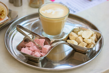 Cappuccino latte macchiato coffee hot drink in a transparent cup, glass on a silver round tray. Pink and brown cane burst sugar in bowls top view. Fresh delicious drink. Sugar tongs. Serving Platter.