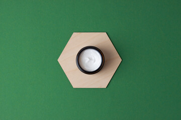 Jar of cosmetic cream with a wooden hexagon on green background. Flat lay, copy space