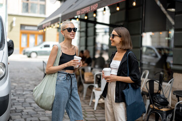 Portrait of a young stylish female couple standing together with a coffee cups outdoors. Street fashion and lifestyle, homosexual relations