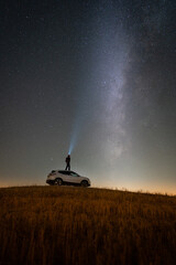 Young man with a headlight standing on a car, looking at the Milky Way