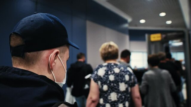 Young Man Wearing Mask And Backpack With Blue Cap Walks To Airplane Along Bridge Among Passengers And Stands In Line