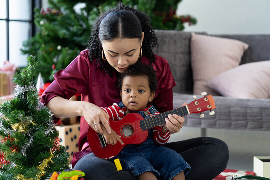 African American mother playing ukulele or small guitar with toddler little boy  on Christmas holiday. African mom playing, having fun, spending time with little boy at home. Merry Christmas