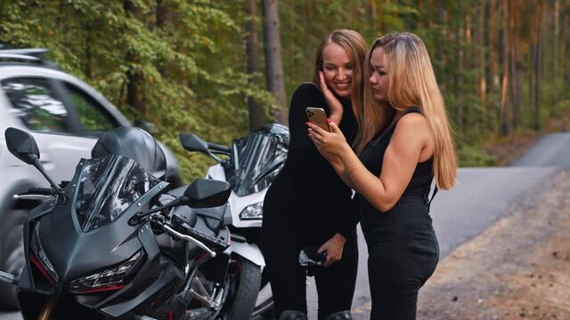 Smiling women friends standing near their motorbikes in the forest and looking at the pictures they made in the phone