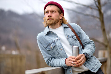 Young caucasian bearded man in nature, looking at side, enjoying the scenery, landscapes. Handsome guy in casual wear traveling alone, holding thermos, looking at interesting sights around him