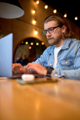 Fototapeta na wymiar hipster lifestyle and creative workspace, caucasian guy working on laptop in coffee shop. bearded man using computer in a cafe, side view shot. copy space. focus on male looking at screen of laptop