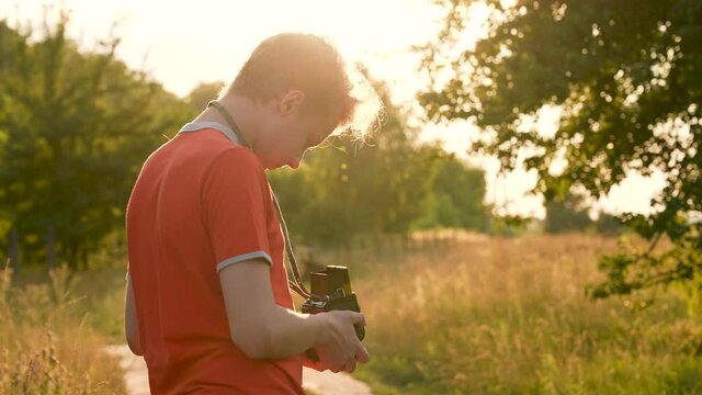 Close-up view of one young white handsome teenage boy using old vintage photo camera while standing outdoors in sunset scenic countryside landscape background