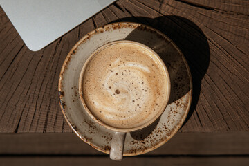Cup of black coffee with milk in beige craft cup on a solid wood oak coffee table with silver laptop. Cozy space for work at home during COVID-19 lock-down period