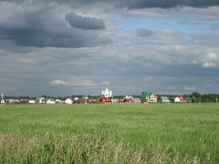 View of a green field overgrown with grass. Behind the field, you can see a village with a towering bell tower of an Orthodox church.