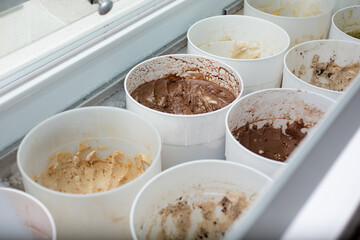 A view looking into a cold storage display at a local ice cream shop, featuring a variety of flavors inside plastic containers.