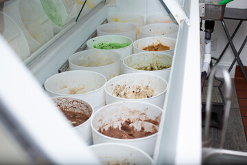 A view looking into a cold storage display at a local ice cream shop, featuring a variety of flavors inside plastic containers.