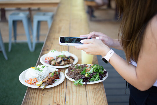 A view of a woman using a cell phone to take food photos outside a local Asian restaurant.