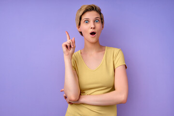 Portrait Of Cheerful Female In Yellow Casual T-shirt Having Great Brilliant Idea, Standing With Wide Opened Mouth, Raising Finger Up, Thinking Creating New Plan, Isolated On Purple Background