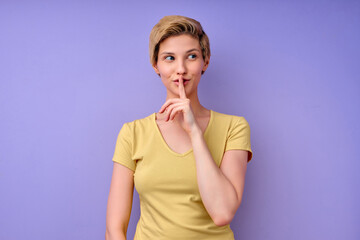Studio shot of quiet attractive lady keeps fore finger over lips, makes silence gesture, gossips with friend, wears yellow t-shirt, says hush, isolated over purple studio wall, looking at side
