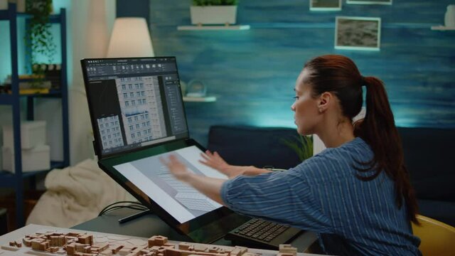 Young engineer using technology for construction design in office. Woman working as architect with building model on touch screen and maquette. Businesswoman analyzing blueprint plan.