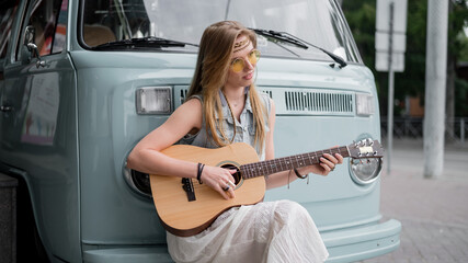 Dreamy woman sits by the camper. A hippie girl holding a guitar and leaning on a classic travel van.