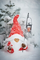 Garden gnome standing under a snow-covered fir. Holds a lantern with a Christmas candle.